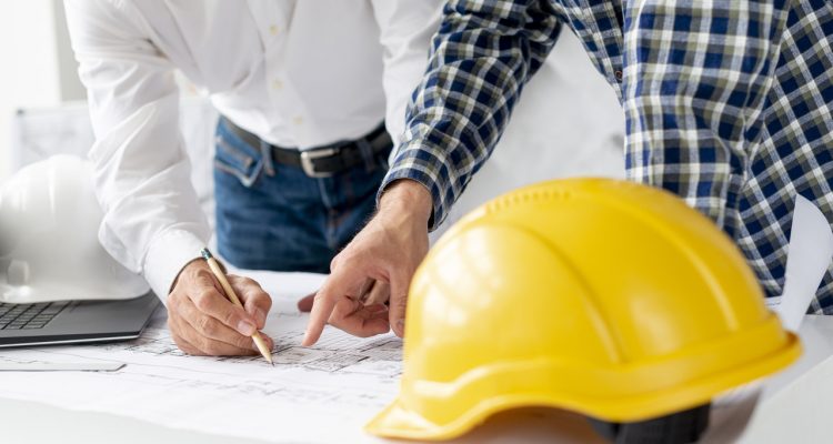 dois homens apontando para projeto e capacete amarelo na mesa